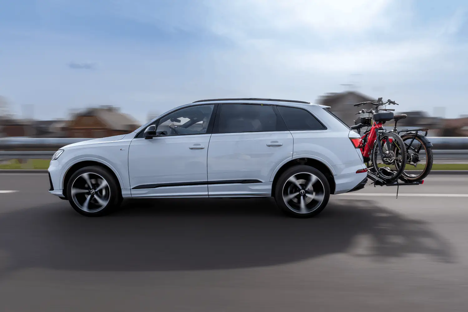 Zdjęcie w ruchu z efektem rozmycia tła (panning) przedstawiające biały SUV marki Audi jadący po asfaltowej drodze. Sylwetka samochodu widziana z boku jest ostra, podczas gdy tło oraz duże, pięcioramienne felgi są dynamicznie rozmyte. Na haku holowniczym z tyłu auta zamontowany jest czarny bagażnik platformowy, na którym przewożony jest jeden rower z czarno-czerwoną ramą. W mocno rozmytym tle widać zarysy barier energochłonnych oraz budynków mieszkalnych pod jasnobłękitnym niebem z delikatnymi chmurami.