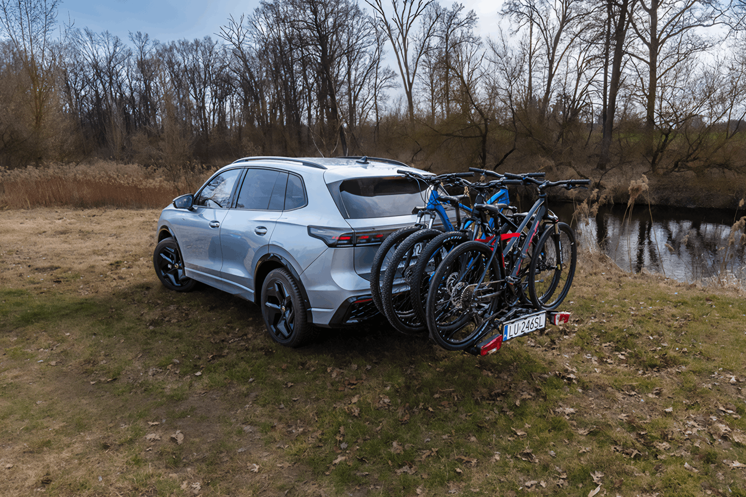 Srebrny SUV zaparkowany tyłem na suchym, trawiastym brzegu rzeki lub stawu. Na haku holowniczym pojazdu zamontowana jest czarna platforma rowerowa, na której przewożone są trzy lub cztery rowery górskie (widoczne są ramy w kolorze niebieskim oraz czarnym z czerwonymi elementami). Tył samochodu jest skierowany lekko w prawą stronę, ukazując polską tablicę rejestracyjną „LU 246SL” oraz czerwone światła pozycyjne bagażnika. Po prawej stronie kadru płynie spokojna woda, w której odbijają się zarośla. W tle rozciąga się linia szarych, pozbawionych liści drzew pod jasnym, lekko zachmurzonym niebem.