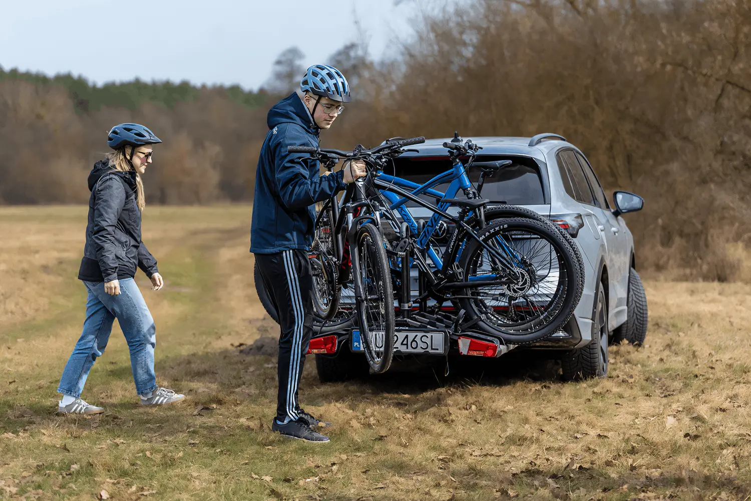 Mężczyzna i kobieta na suchej, trawiastej polanie na tle bezlistnych, brązowych drzew wczesną wiosną lub jesienią. Po prawej stronie stoi srebrny SUV z zamontowanym na haku holowniczym platformowym bagażnikiem rowerowym. Mężczyzna w sportowym ubraniu (ciemnoniebieska kurtka z kapturem, czarne spodnie dresowe z trzema białymi paskami) oraz w jasnoniebieskim kasku mocuje na bagażniku jaskrawoniebieski rower górski. Z lewej strony w jego kierunku idzie kobieta w ciemnej kurtce, jeansach, jasnych butach sportowych i ciemnym kasku. Na bagażniku samochodowym wyraźnie widać białą polską tablicę rejestracyjną z numerem „LU 246SL”.
