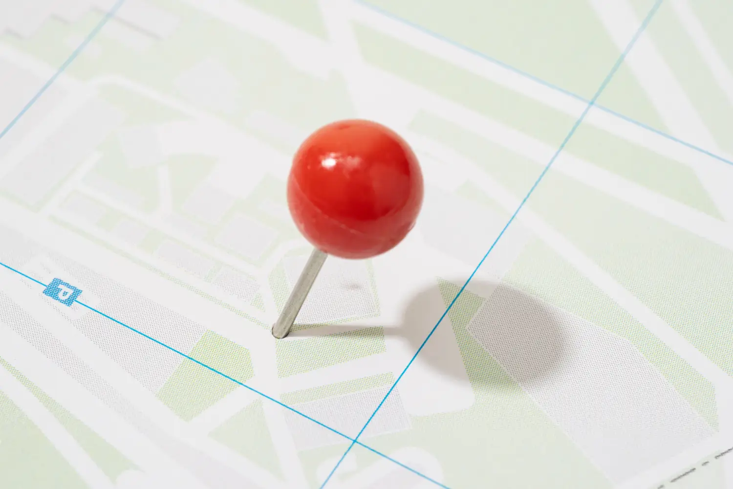 Zdjęcie makro przedstawiające dużą pinezkę z okrągłą, błyszczącą, czerwoną główką wbitą pod skosem w papierową mapę. Mapa ma minimalistyczną szatę graficzną w jasnych barwach – dominują odcienie bieli i pastelowej zieleni, z cienkimi niebieskimi liniami wyznaczającymi siatkę geograficzną lub ulice. W pobliżu metalowej nóżki pinezki widoczny jest mały, niebieski kwadratowy symbol parkingu z literą „P”. Pinezka rzuca wyraźny, miękki cień w prawą stronę na powierzchnię mapy.