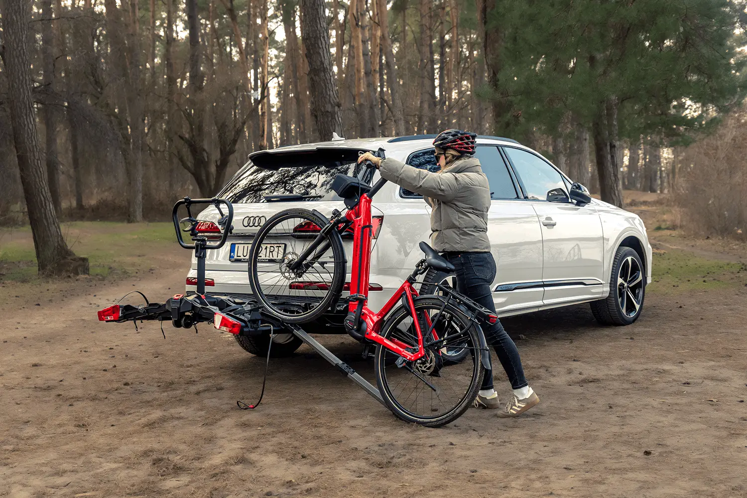 Kobieta w szarej kurtce puchowej, ciemnych jeansach i kasku ochronnym wprowadza czerwony rower elektryczny na czarną platformę rowerową zamontowaną na haku białego SUV-a marki Audi. Koła roweru są prowadzone wzdłuż specjalnej rampy załadunkowej, która łączy podłoże z rynną bagażnika. Sytuacja ma miejsce na piaszczystym terenie w otoczeniu rzadkiego lasu sosnowego, przy naturalnym, popołudniowym świetle.