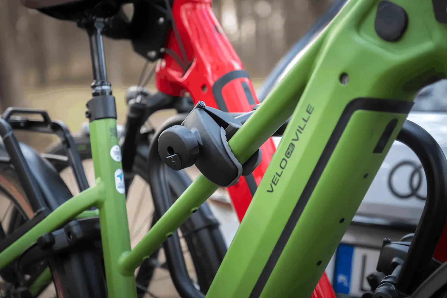 Bliskie ujęcie detalu technicznego platformy rowerowej. Czarna, solidna klamra mocująca z szarym, gumowym wykończeniem od wewnątrz jest mocno zaciśnięta na profilu jasnozielonej ramy roweru elektrycznego marki VELO DE VILLE. Konstrukcja uchwytu wyposażona jest w zamek zabezpieczający na kluczyk. W mocno rozmytym tle widać fragment konstrukcji bagażnika oraz ramę drugiego roweru w intensywnym, czerwonym kolorze.