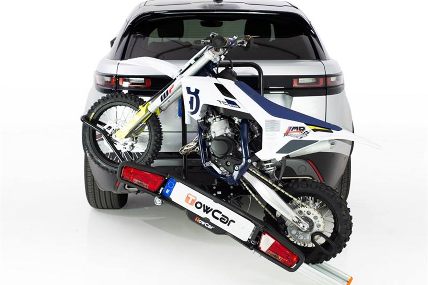 Srebrny samochód osobowy z zamontowanym na haku bagażnikiem platformowym TowCar w pozycji odchylonej, ułatwiającej załadunek biało-niebieskiego motocykla crossowego marki Husqvarna.