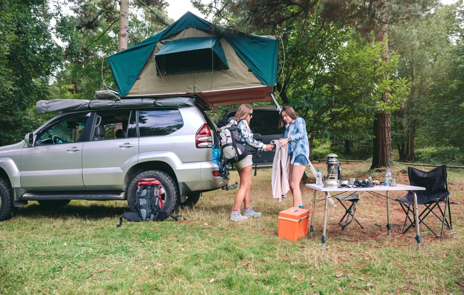  Srebrny SUV zaparkowany na leśnej polanie, z rozłożonym na dachu ciemnozielonym namiotem turystycznym. Dwie młode kobiety przy otwartym bagażniku składają koc i pakują sprzęt. Na pierwszym planie stoi składany stolik kempingowy z lampą naftową, lornetką i butelkami wody oraz pomarańczowa lodówka turystyczna.