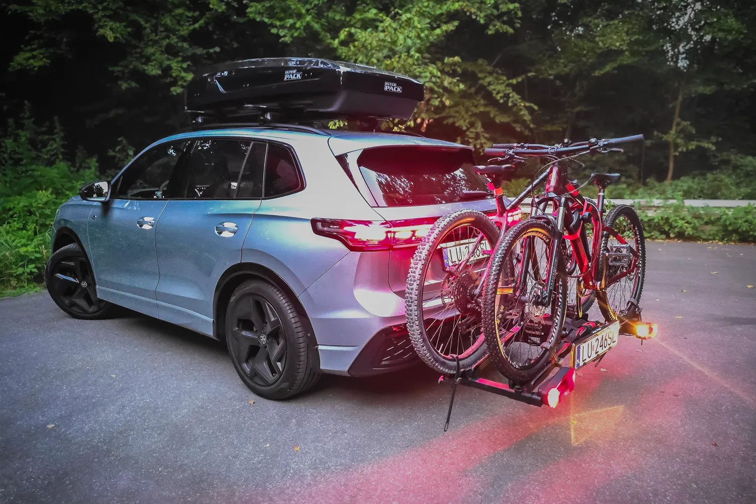 Srebrny SUV Volkswagen Tiguan z czarnymi felgami, zaparkowany na asfaltowej drodze o zmierzchu. Na srebrnych relingach samochodu zamontowany jest duży, aerodynamiczny box dachowy Inter Pack w kolorze głębokiego, błyszczącego czarnego lakieru. Z tyłu pojazdu, na haku, znajduje się platforma rowerowa z podświetloną tablicą rejestracyjną i włączonymi czerwonymi światłami pozycyjnymi. Na platformie przypięte są dwa rowery górskie MTB o czarno-czerwonych ramach. W tle za samochodem rozciąga się gęsta, ciemna ściana lasu, a cała kompozycja ma wieczorny, podróżniczy klimat.