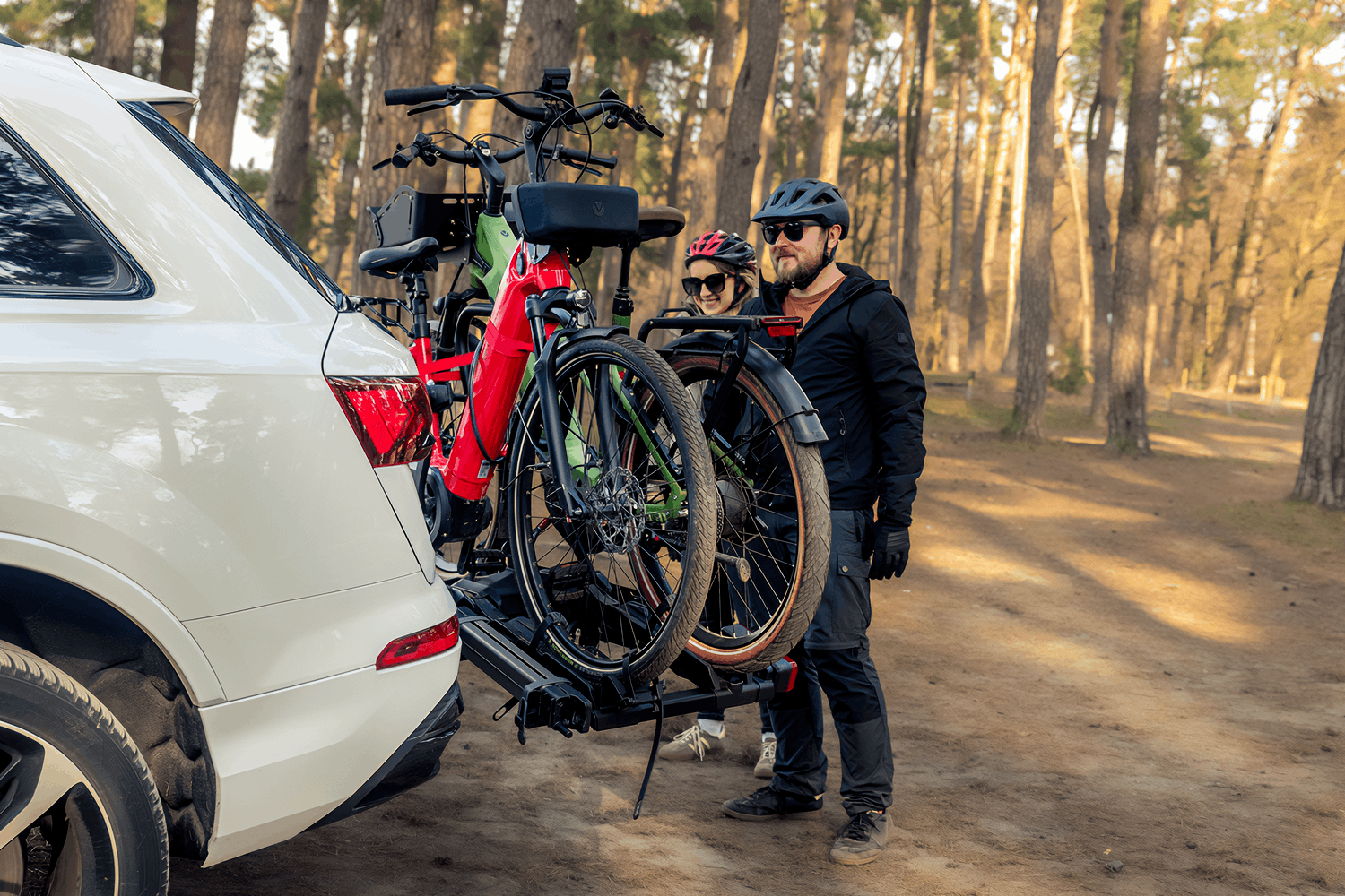 Bliski kadr na prawy tył białego samochodu typu SUV z zamontowaną na haku holowniczym czarną platformą rowerową. Na bagażniku zabezpieczone są pionowo dwa rowery elektryczne; pierwszy z nich ma wyrazistą, matową czerwoną ramę, czarny przedni amortyzator, tarczowe hamulce hydrauliczne oraz podłużną czarną torbę przypiętą do kierownicy. Za kołami rowerów stoi para w kaskach – brodaty mężczyzna w ciemnej kurtce i okularach przygląda się mocowaniu, a obok niego w tle uśmiecha się kobieta w czerwonym kasku. Scena rozgrywa się na piaszczystym podłożu pod lasem, w otoczeniu wysokich sosen w miękkim świetle dnia.