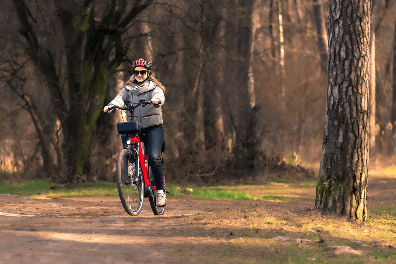 Portret uśmiechniętej kobiety w kasku rowerowym i okularach przeciwsłonecznych, jadącej czerwonym rowerem trekkingowym z torbą na kierownicy. Kobieta jest ubrana w szarą kurtkę puchową i ciepły szalik. Porusza się po ubitej, ziemnej ścieżce wewnątrz jasnego lasu, gdzie wysokie pnie drzew i naturalne światło tworzą malownicze tło z łagodnym efektem bokeh.