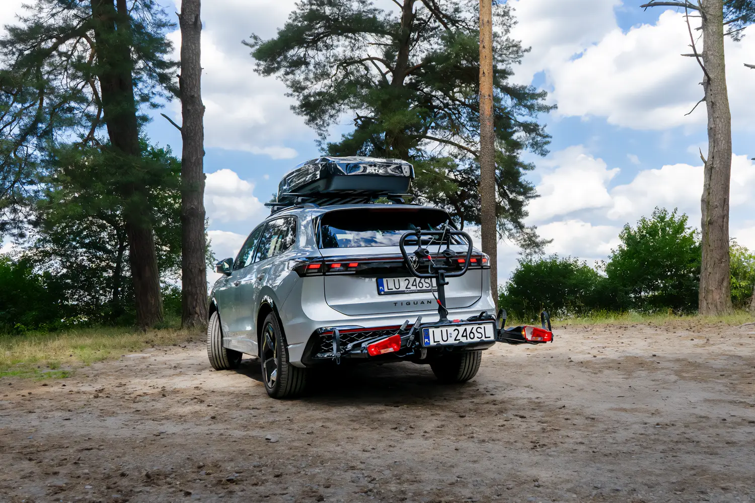 Srebrny SUV marki Volkswagen Tiguan zaparkowany na piaszczystej leśnej drodze w otoczeniu wysokich sosen. Na dachu samochodu zamontowano czarny box dachowy marki Inter Pack, a na haku holowniczym znajduje się złożona platforma rowerowa z widocznymi tylnymi światłami i tablicą rejestracyjną.