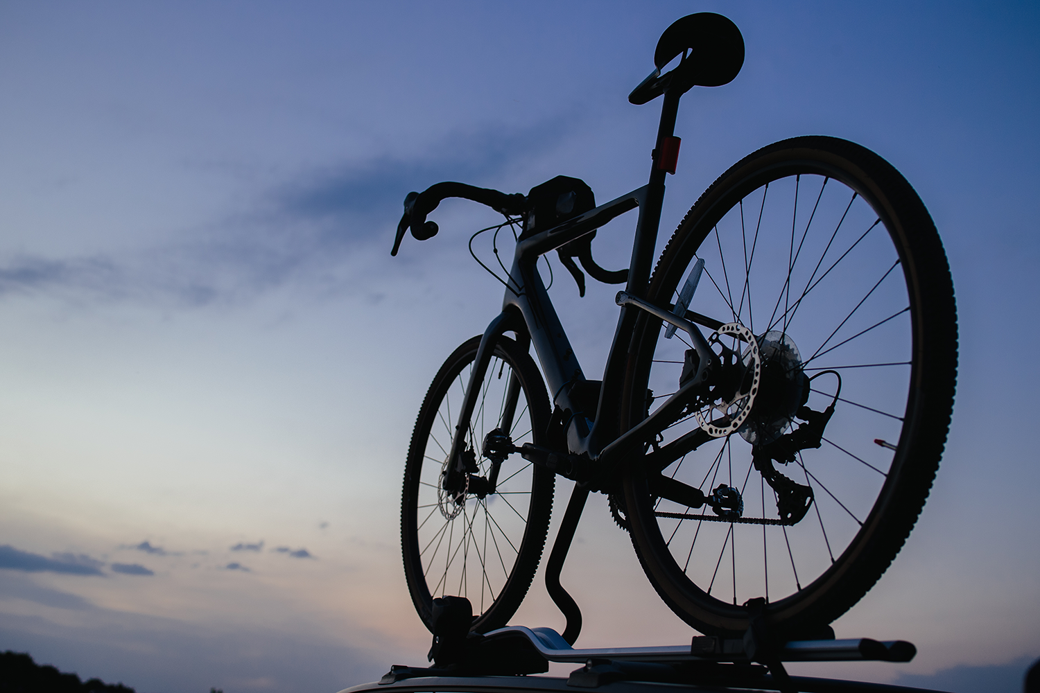 Sylwetka roweru szosowego lub gravelowego z kierownicą typu baranek, zamontowanego na dachowym uchwycie rowerowym, sfotografowana z niskiej perspektywy na tle ciemniejącego nieba o zmierzchu. Widoczne są precyzyjne detale ramy, tarczy hamulcowej, szprych oraz siodełka zarysowane na tle niebiesko-pomarańczowego horyzontu.