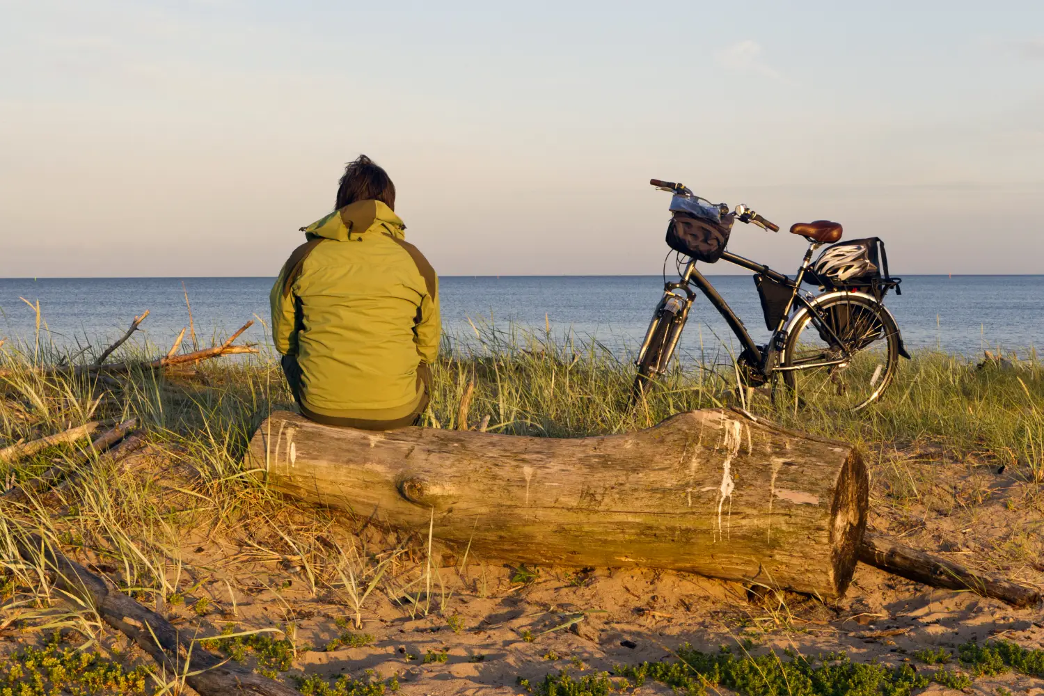 Rowerzysta w zielonej kurtce siedzi tyłem do kadru na drewnianym pniu na piaszczystej plaży, obserwując spokojne morze o złotej godzinie. Obok niego stoi czarny rower turystyczny z torbą na kierownicy, sakwą oraz kaskiem przymocowanym do tylnego bagażnika.