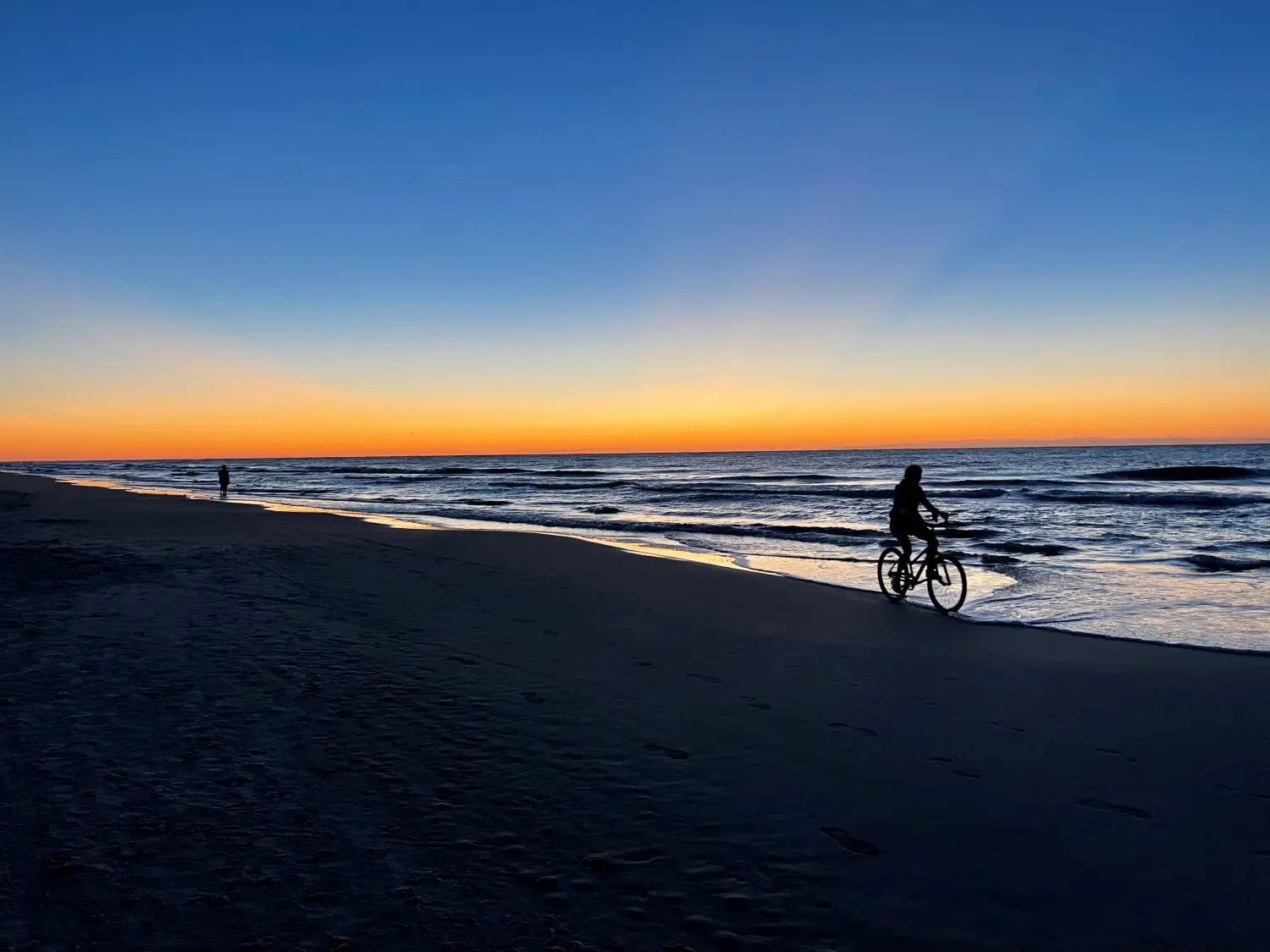 Ciemna sylwetka rowerzysty jadącego brzegiem piaszczystej plaży tuż przy falach morskich podczas spektakularnego zachodu słońca. Niebo na horyzoncie płonie jaskrawym, pomarańczowym światłem, przechodzącym wyżej w głęboki błękit.