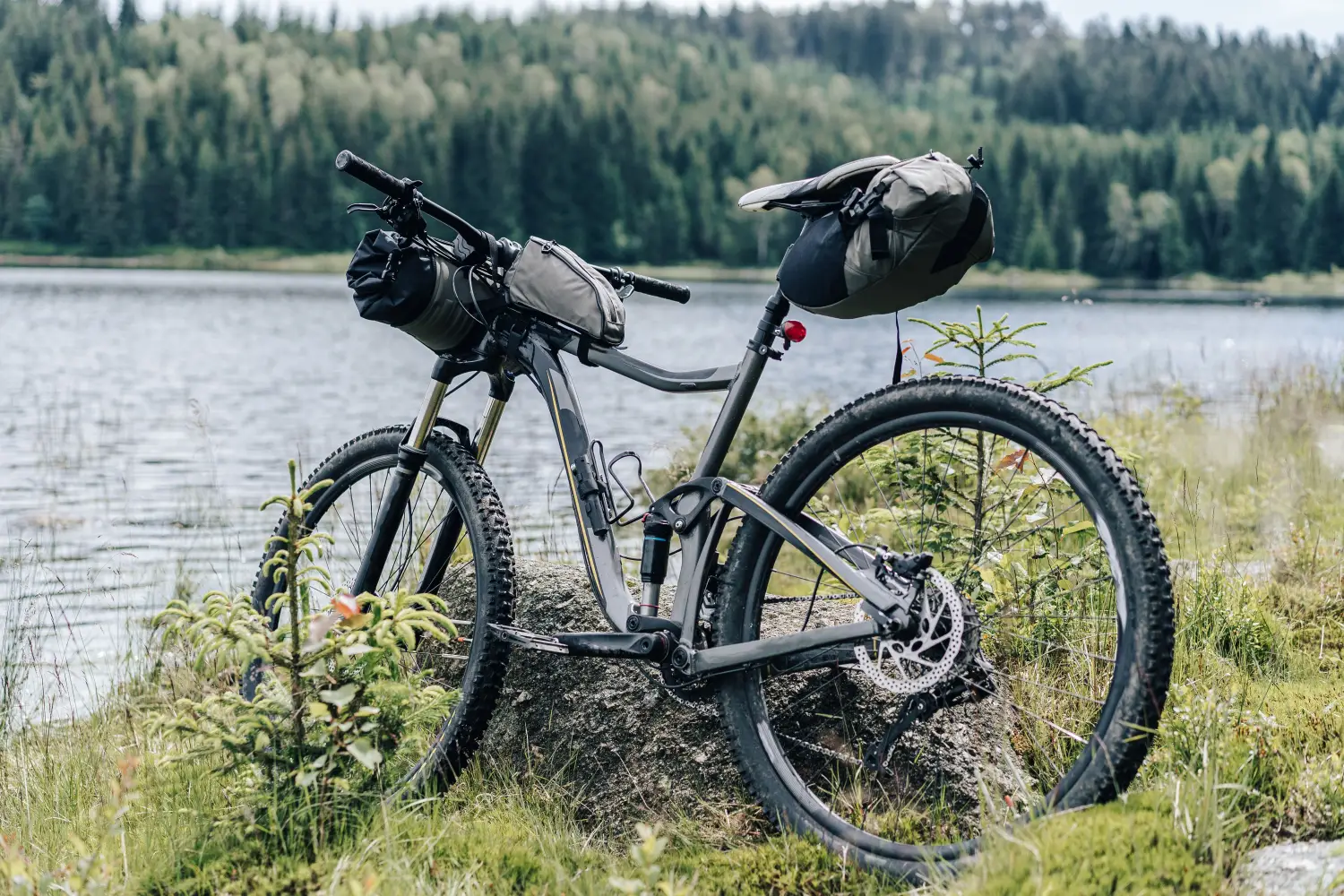 Szary rower górski z pełną amortyzacją (full suspension), oparty o duży głaz na trawiastym brzegu jeziora. Rower wyposażony jest w komplet toreb do bikepackingu: rolkę na kierownicy, małą torbę na górnej rurze ramy oraz dużą oliwkowo-czarną torbę podsiodłową. W tle rozciąga się tafla wody oraz gęsty, iglasty las pod zachmurzonym niebem.
