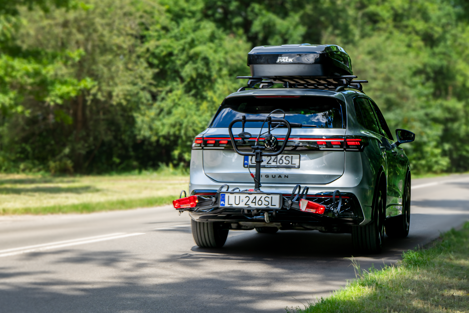 Jasnosrebrny Volkswagen Tiguan SUV jadący po asfaltowej drodze w otoczeniu zielonego lasu. Samochód wyposażony jest w dwa systemy transportowe markowane przez Inter Pack: na dachu zamontowano czarny, zamykany box dachowy ustawiony na aluminiowym bagażniku bazowym, natomiast z tyłu, na haku holowniczym, znajduje się złożona, pusta platforma rowerowa Inter Pack z podświetloną tablicą rejestracyjną LU 246SL, tylnymi lampami i pionowym pałąkiem mocującym.