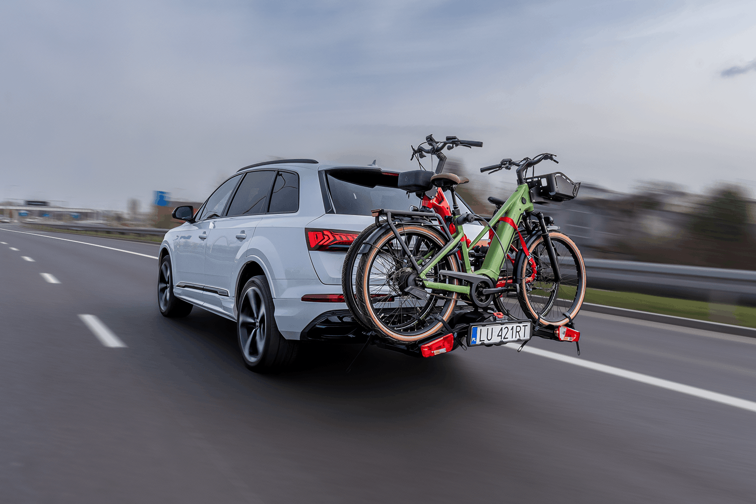 Srebrne Audi Q7 w wersji SUV jadące po wielopasmowej drodze szybkiego ruchu z zamontowaną na haku holowniczym platformą rowerową. Na bagażniku transportowane są dwa rowery elektryczne: zielony na pierwszym planie oraz czerwony za nim. Bagażnik wyposażony jest we własny panel oświetleniowy, czerwone światła pozycyjne oraz dodatkową polską tablicę rejestracyjną o numerze LU 421RT. Pojazd znajduje się w ruchu na tle barier ochronnych i nieba.