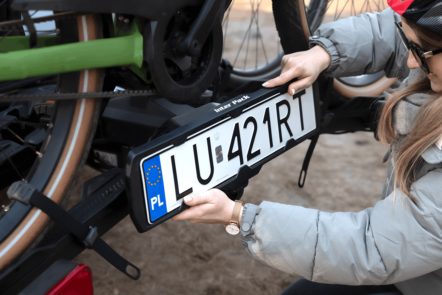 Zbliżenie z bliska na dłonie kobiety w szarej kurtce zimowej, która montuje polską tablicę rejestracyjną o numerze LU 421RT w czarnej ramce z napisem Inter Pack. Ramka zamocowana jest w dolnej części platformy rowerowej, tuż pod kołami zainstalowanego roweru elektrycznego o zielonej ramie i jasnych oponach. Kobieta trzyma ramkę od góry i od dołu, precyzyjnie wpinając tablicę w zatrzaski bagażnika.