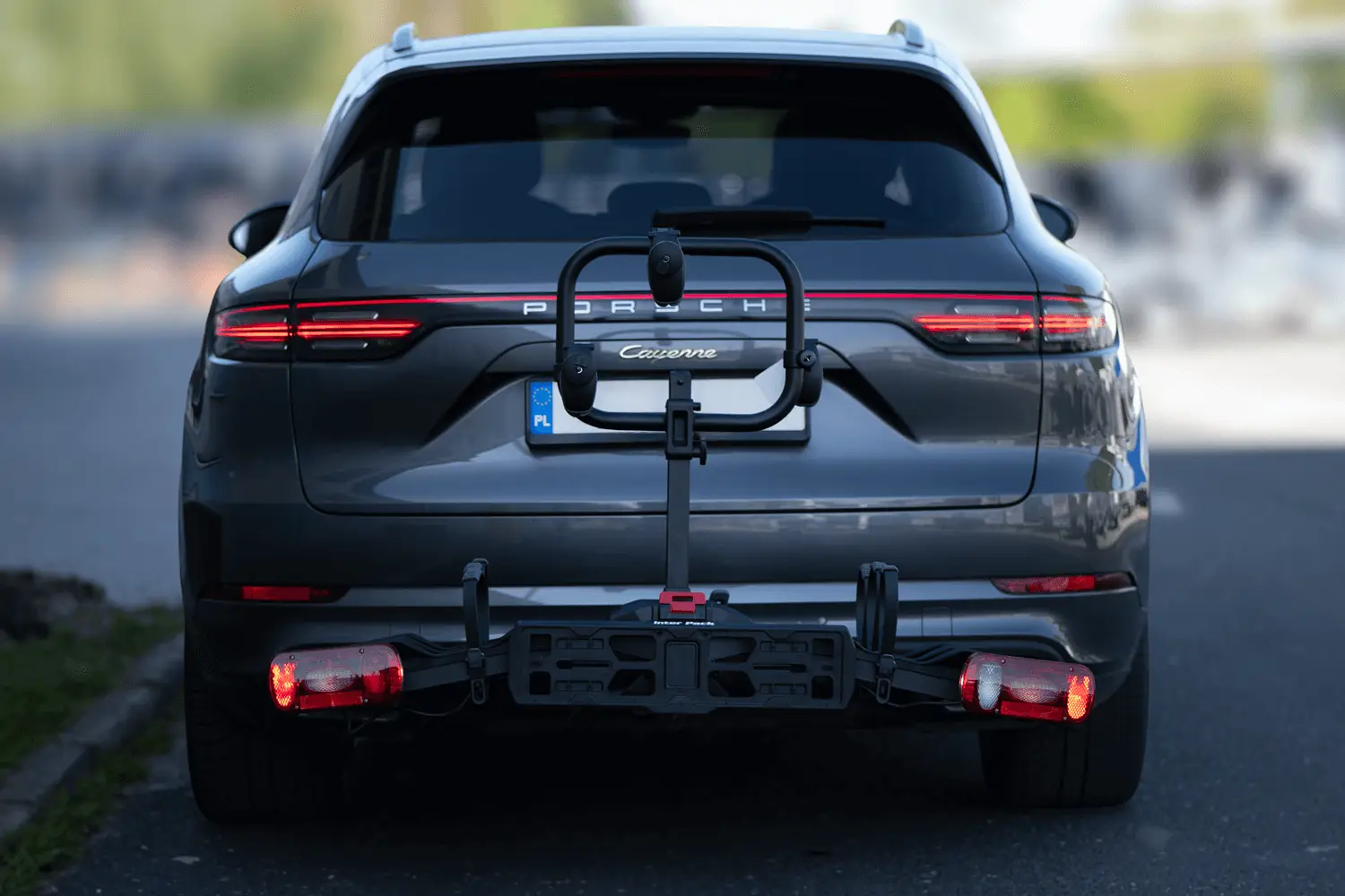 Widok z tyłu na szary samochód SUV marki Porsche Cayenne zaparkowany na drodze, z zamontowanym na haku holowniczym pustym bagażnikiem rowerowym.