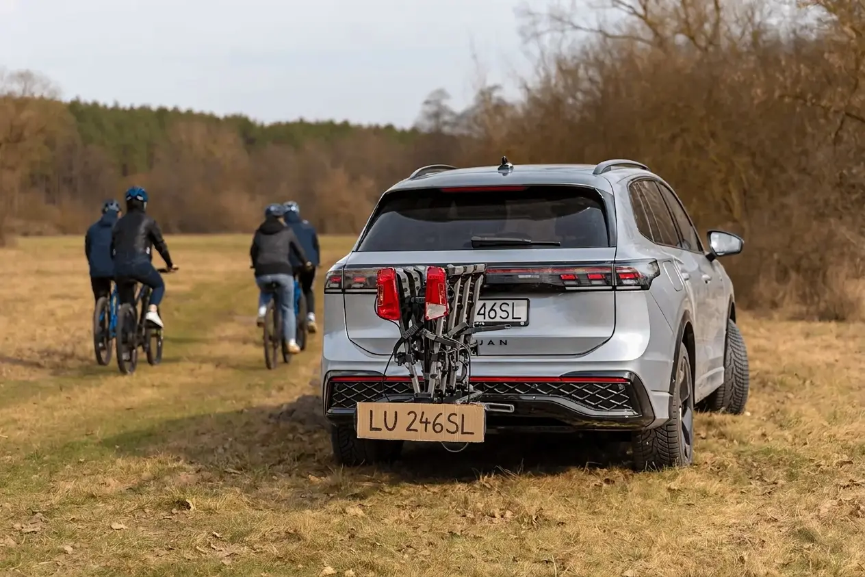  Srebrny samochód SUV z zamontowanym na haku złożonym bagażnikiem rowerowym stoi na polnej drodze, podczas gdy w tle cztery osoby jadą na rowerach.