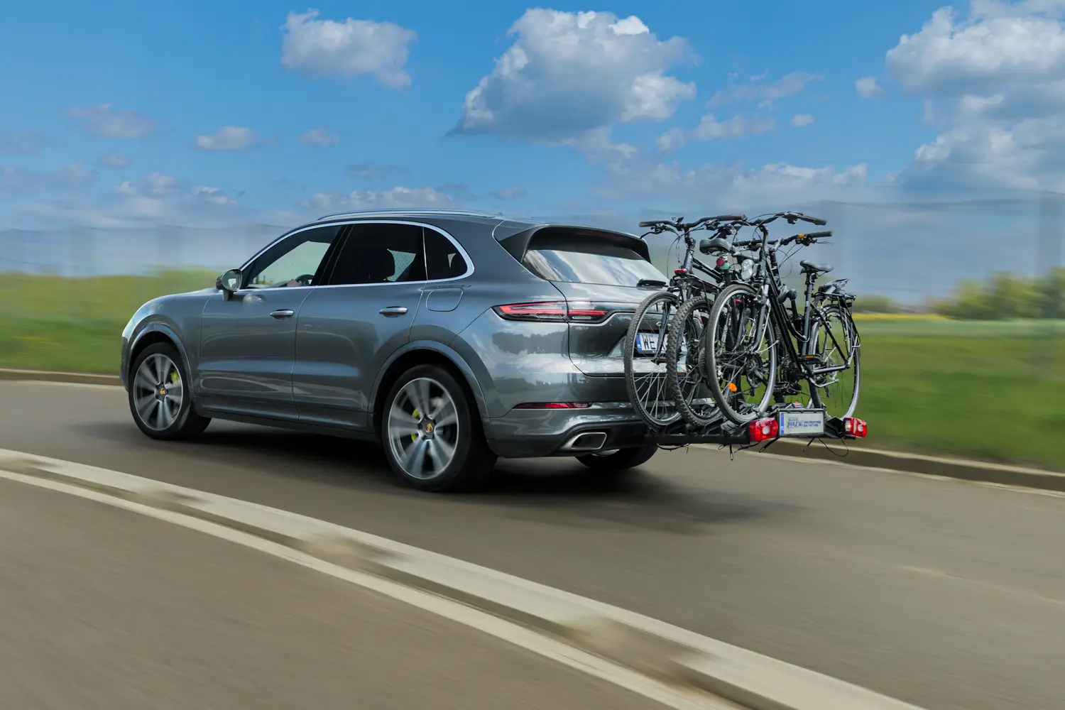 Ciemnoszary SUV marki Porsche Cayenne w ruchu na dwupasmowej drodze, widziany z profilu od tyłu. Na haku holowniczym samochodu zamontowano platformę rowerową Inter Pack ze światłami tylnymi oraz tablicą rejestracyjną. Na platformie przewożone są trzy czarne rowery miejskie i trekkingowe. Pojazd jedzie wzdłuż pasa zieleni na tle jasnoniebieskiego nieba z białymi chmurami.