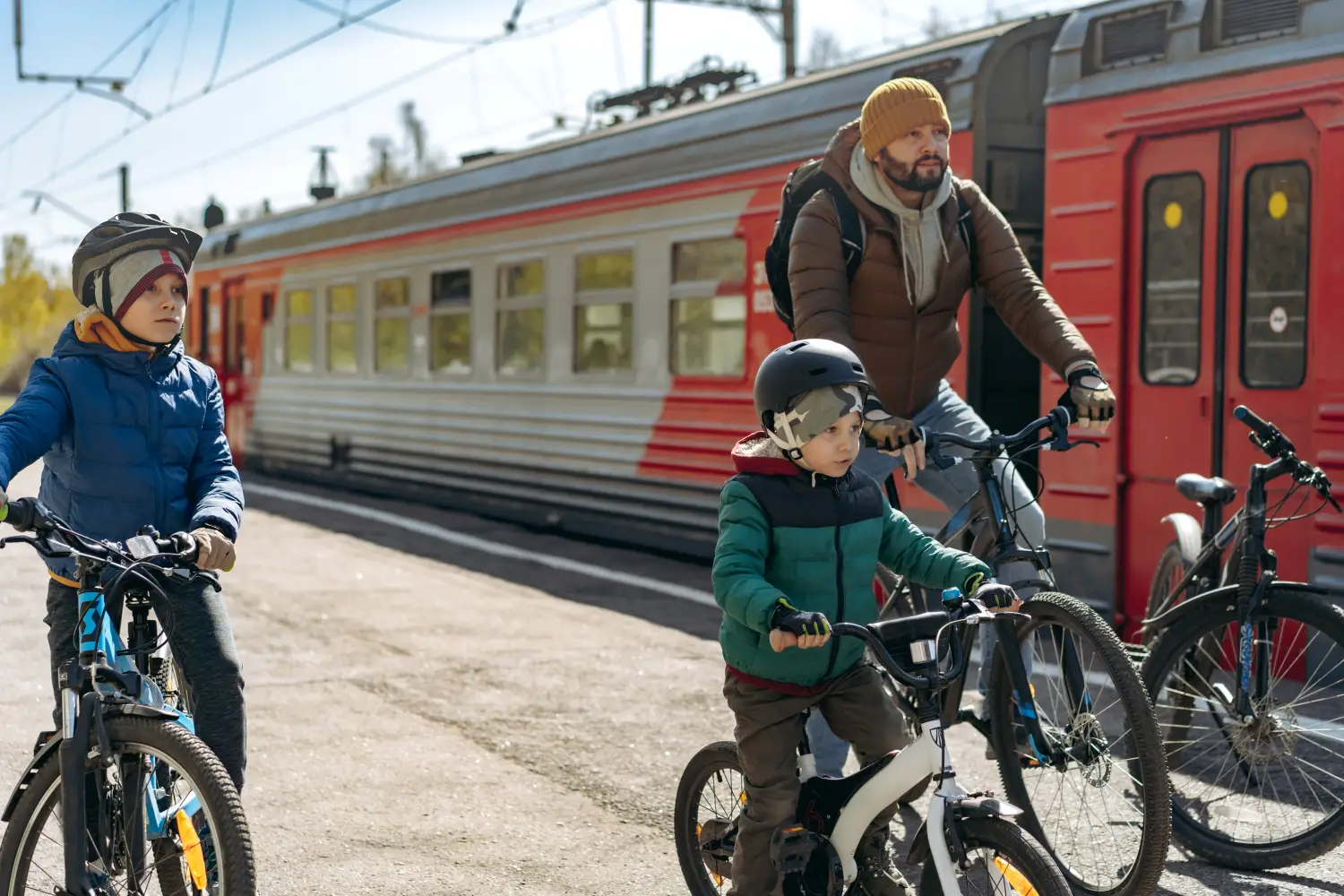 Mężczyzna w brązowej kurtce i żółtej czapce oraz dwójka małych chłopców w kaskach rowerowych stojący ze swoimi rowerami na peronie przy czerwono-szarym pociągu osobowym. Chłopiec na pierwszym planie po lewej jedzie na niebieskim rowerze górzystym, młodszy syn na środku stoi na małym białym rowerku, a ojciec z tyłu trzyma czarny rower trekkingowy. Rodzina wyjeżdża na wycieczkę rowerową w słoneczny dzień.