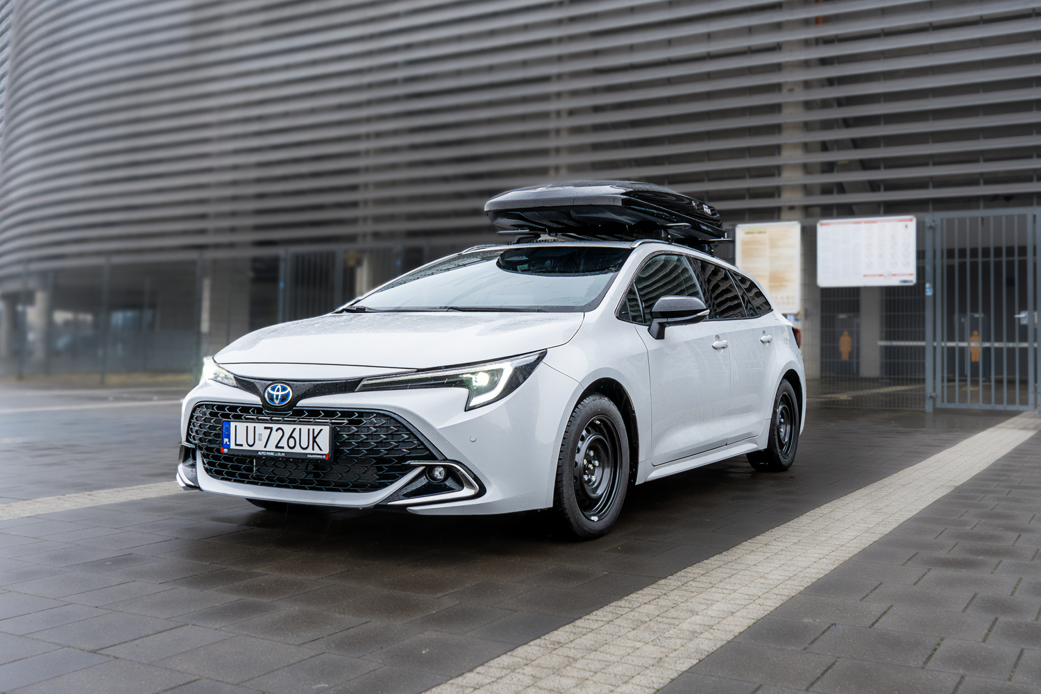 Biała Toyota Corolla stojąca na tle ściany stadionu. Na samochodzie zamontowany czarny box dachowy