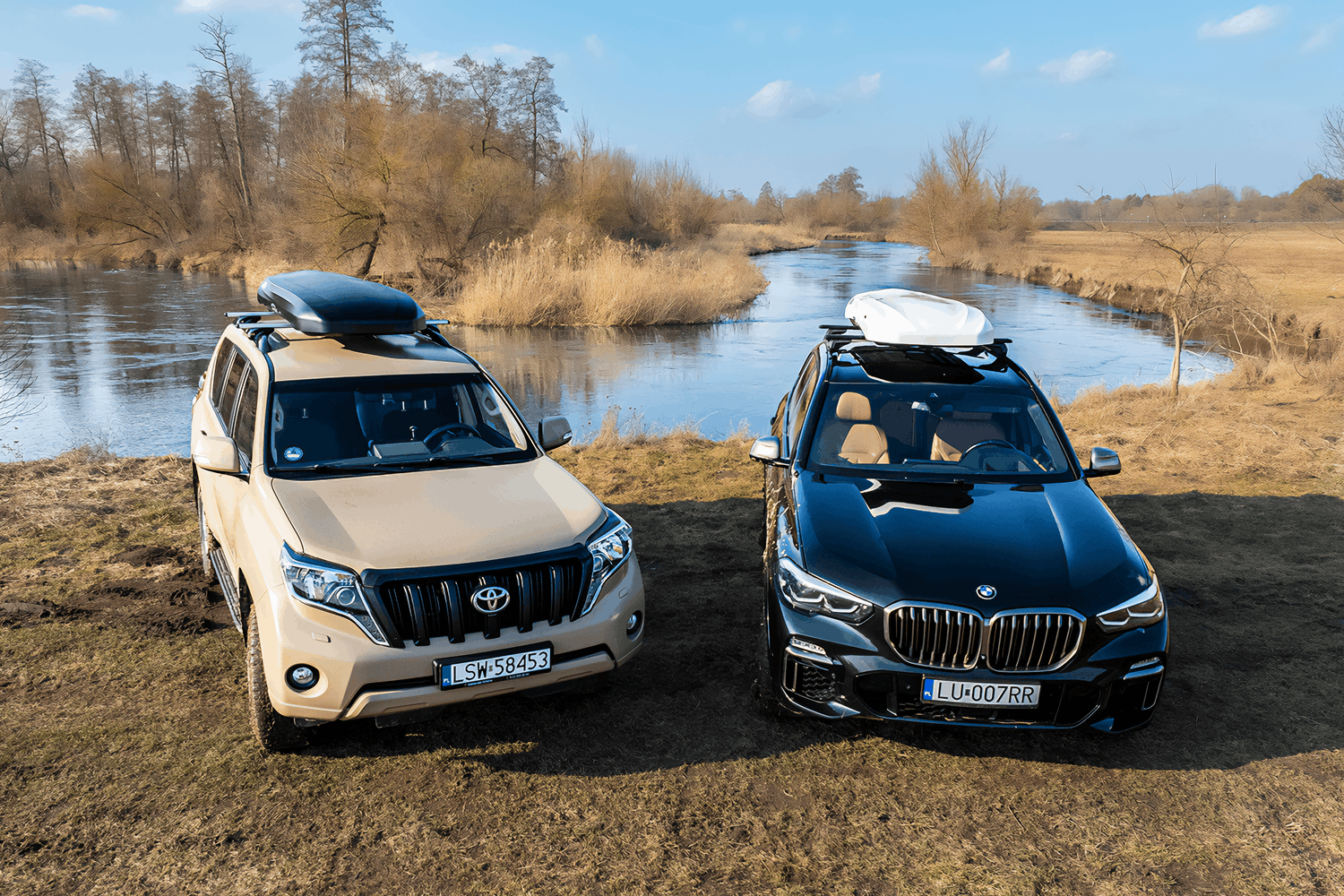 Dwa auta stojące nad brzegiem wąskiej rzeki. Po prawej czarne auto marki BMW z biały boxem na dachu. Po lewej beżowa Toyota z czarnym boxem. W tle krajobraz polany i lasu.