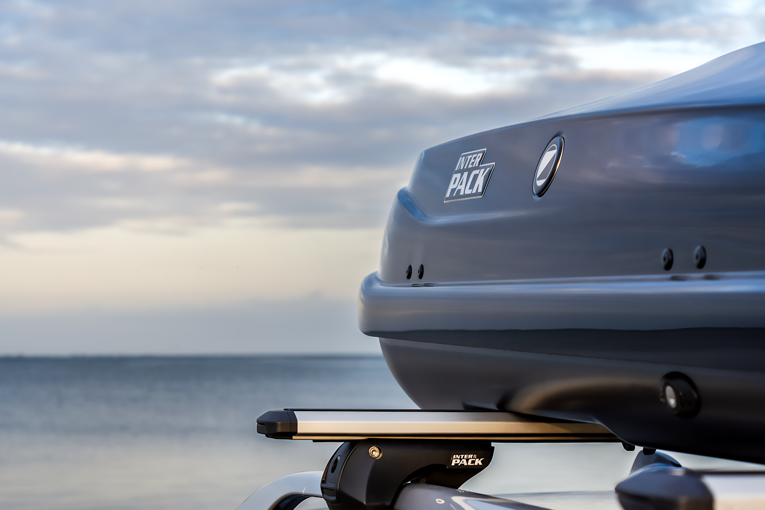 Zbliżenie na czarny box dachowy z logo Inter Pack zamontowany na srebrnych belkach bazowych również z logo. W tle krajobraz morza.