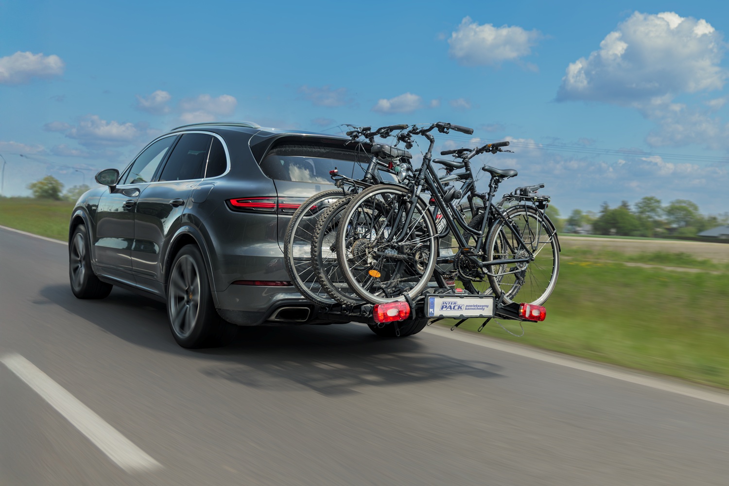 Srebrne Porsche Cayenne z zamontowaną platformą rowerową. Przymocowane do niej 3 rowery. Auto w trakcie jazdy w słoneczny dzień po asfalcie. W tle pola oraz las.