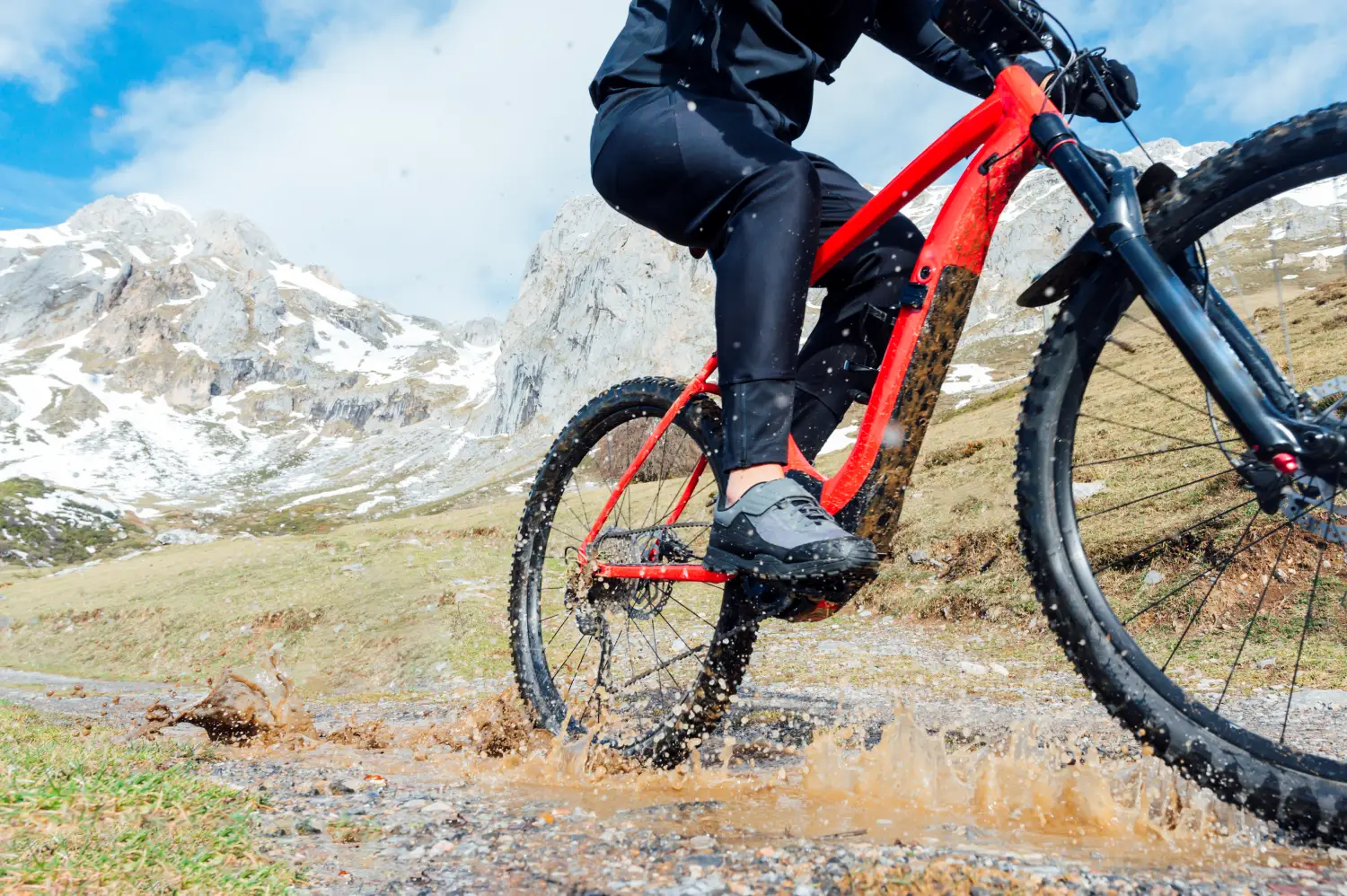 Rowerzysta na czerwonym rowerze górskim dynamicznie przejeżdża przez kałużę, wzlapując błoto i wodę na tle ośnieżonych szczytów górskich.