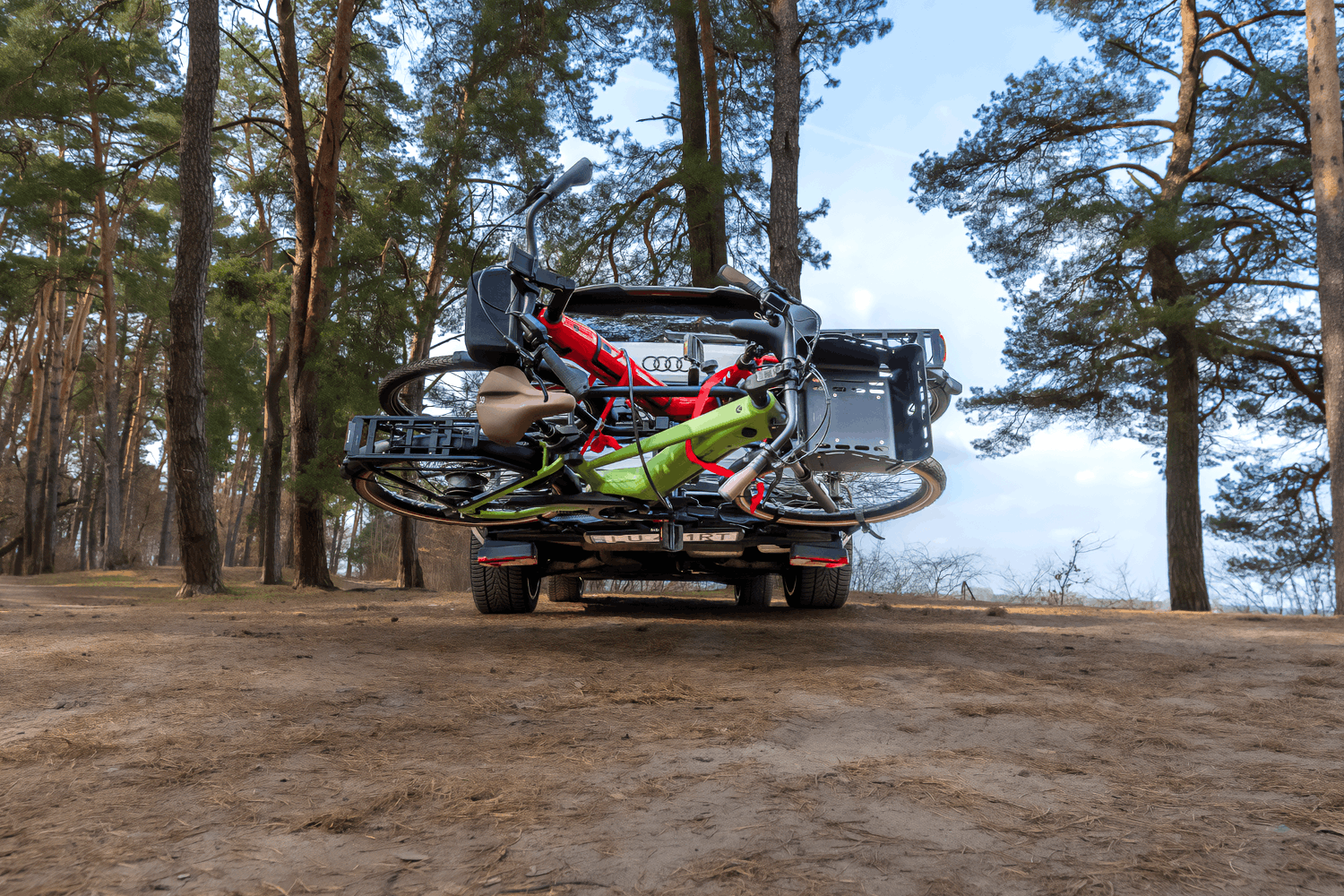 Las, jasny dzień. Na niewielkiej polance między drzewami stoi srebrny SUV Audi. Widok na tył auta z dołu. Na haku holowniczym zamontowana platforma rowerowa z dwoma rowerami miejskimi - czerwonym oraz zielonym.