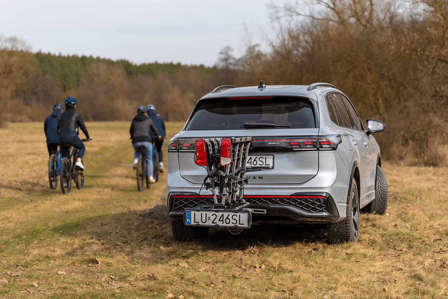 Tył srebrnego SUVa Volkswagena z zamontowaną na haku platformą rowerową. Platforma jest złożona do pionu. Auto stoi po prawej stronie kadru, w tle jesienny las. Po lewej stronie kadru 4 osoby na rowerach, odjeżdżają wgłąb kadru, plecami do obiektywu. Każdy z nich w kasku, czarnej kurtce oraz jeansach.