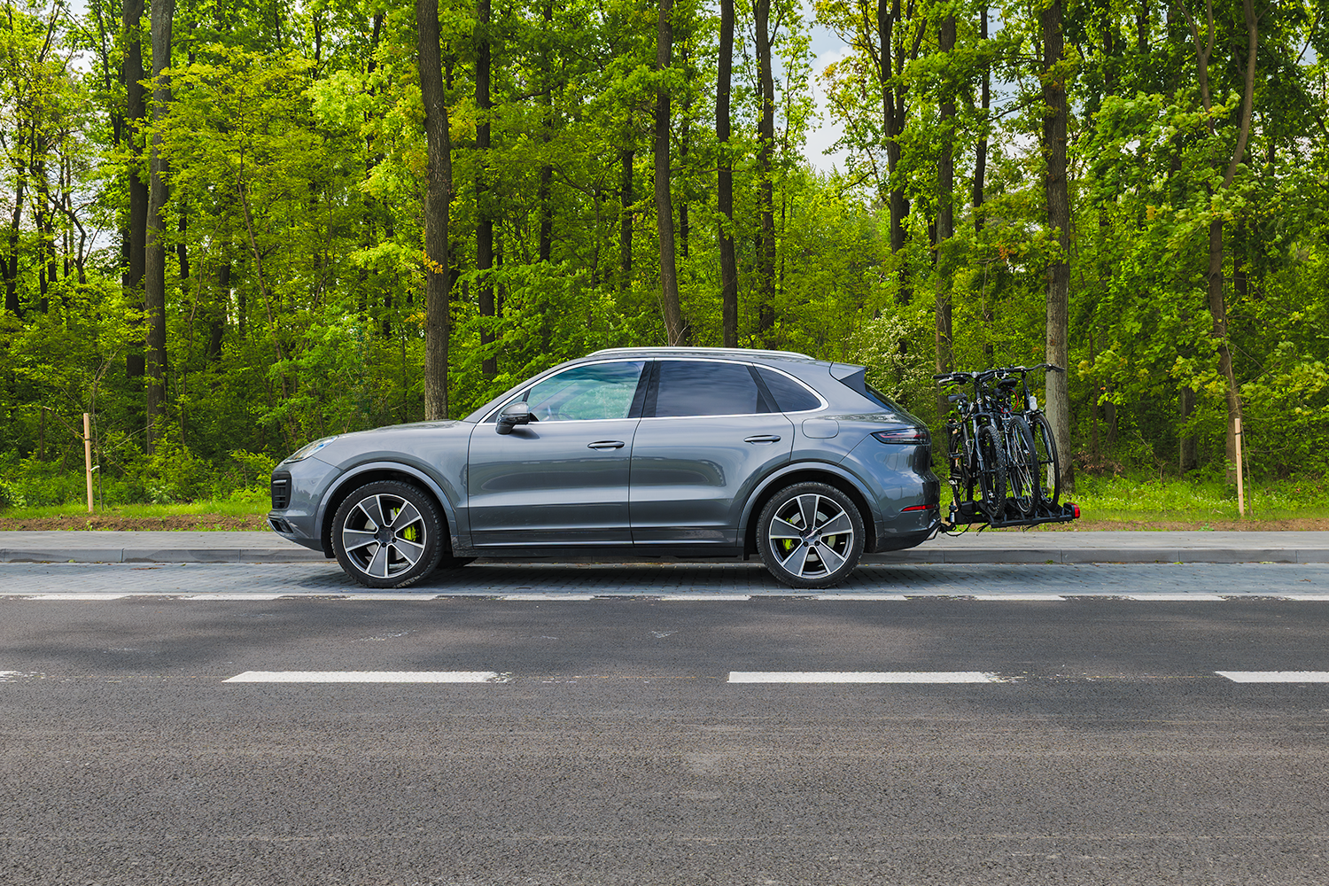 Dwupasmowa droga asfaltowa z poboczem widziana od boku, w tle gęsty las. Jasny, pogodny dzień. Na poboczu srebrny SUV Porsche z zamontowanym na haku holowniczym bagażnikiem rowerowym. Na platformie 3 rowery.