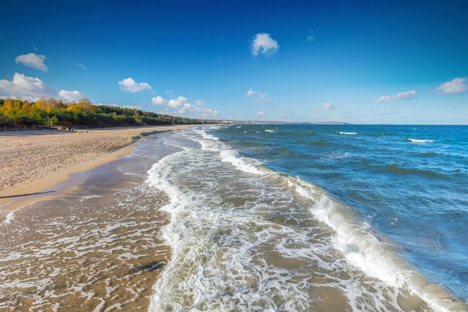 Piaszczysta plaża nad morzem z białymi falami uderzającymi o brzeg pod błękitnym niebem z lekkimi chmurami.
