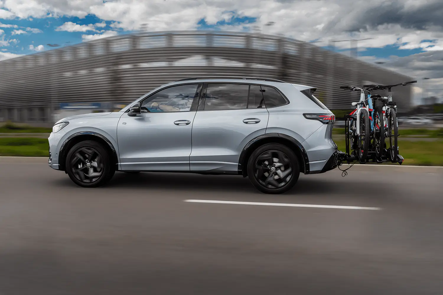  Srebrny SUV w ruchu na drodze, z zamontowanym na haku holowniczym bagażnikiem rowerowym przewożącym rowery.