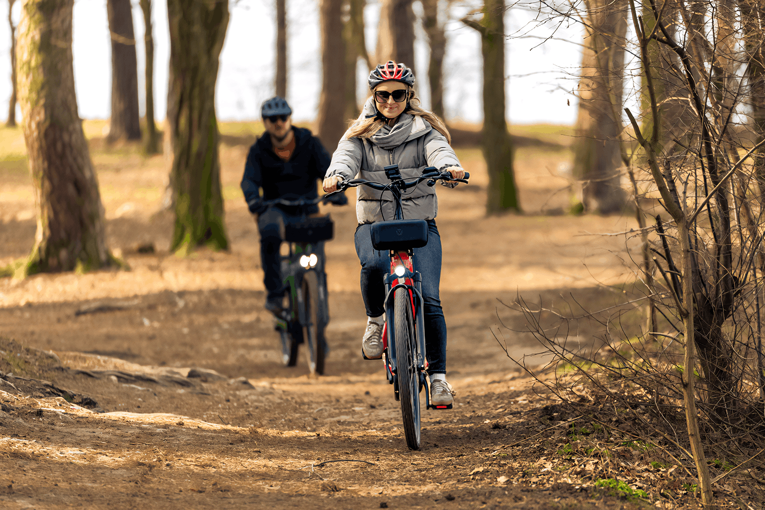Krajobraz brązowego, jesiennego lasu. Leśną ścieżką jadą dwie osoby na rowerach. Z przodu kobieta z blond włosami, okularami przeciwsłonecznymi, szarą kurtką i jeansami na czerwonym rowerze. W tle na czarnym rowerze mężczyzna, również w okularach przeciwsłonecznych oraz ubrany w czarną kurtkę i spodnie.