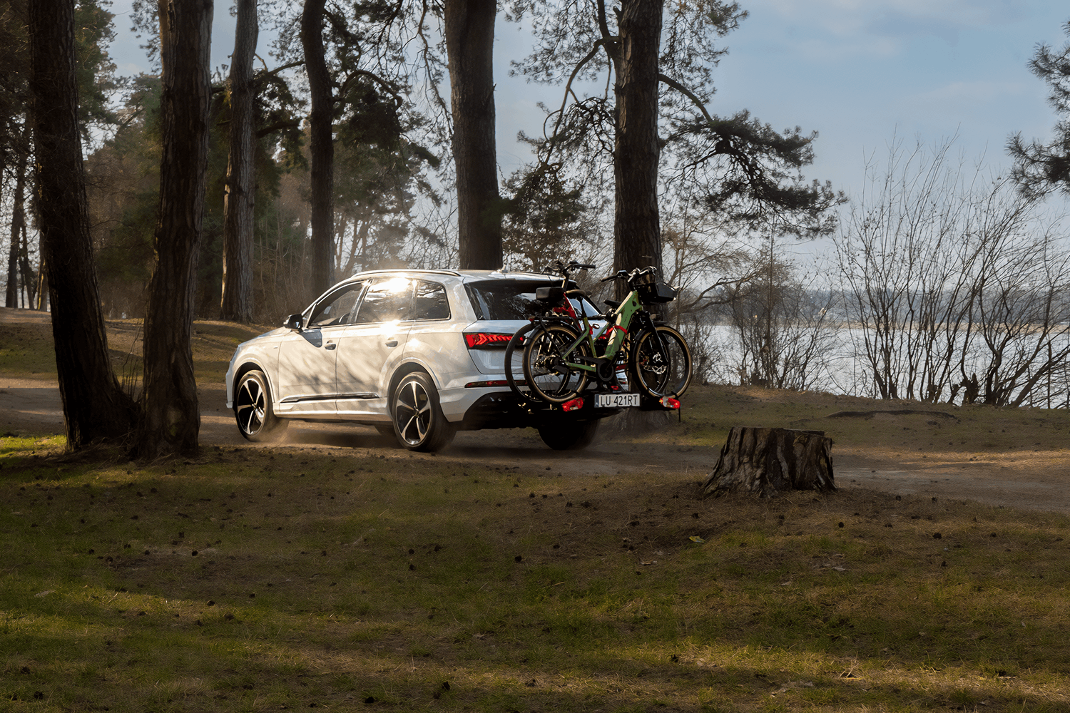 Biały SUV z zamontowaną platformą rowerową i dwoma rowerami. Widok z daleka, krajobraz lasu w trakcie zachodu słońca. W tle widoczny kawałek zalewu