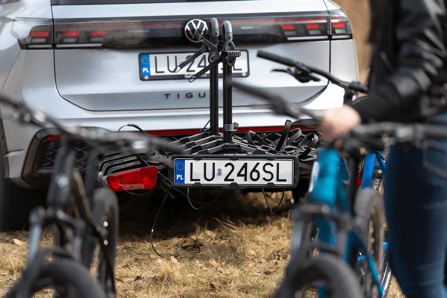 Na pierwszym planie widoczne dwa rozmazane rowery. Po lewej stronie stojący na stopce czarny rower, po prawej niebieski, który jest podtrzymywany przez osobę w czarnej, skórzanej kurtce. Kadr nie obejmuje głowy postaci. Ostrość na stojący w tle srebrny SUV Volkswagena z zamontowaną czarną platformą rowerową na hak holowniczy.
