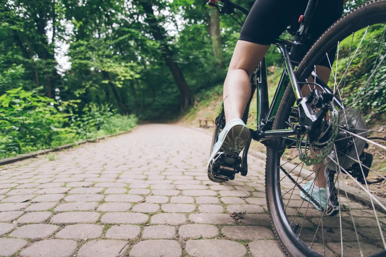 Zbliżenie na dolną część roweru stojącego na brukowanej drodze biegnącej pod górkę. Na lewym pedale roweru noga rowerzysty. Białe buty i krótkie, sportowe spodenki. Wokół brukowanej drogi widoczny las.
