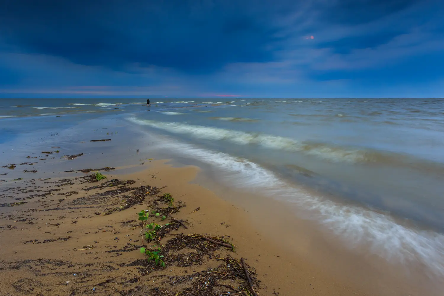 Krajobraz nadmorski o zmierzchu lub w pochmurny dzień, z piaszczystą plażą, delikatnymi falami uderzającymi o brzeg oraz ciemnym, burzowym niebem.