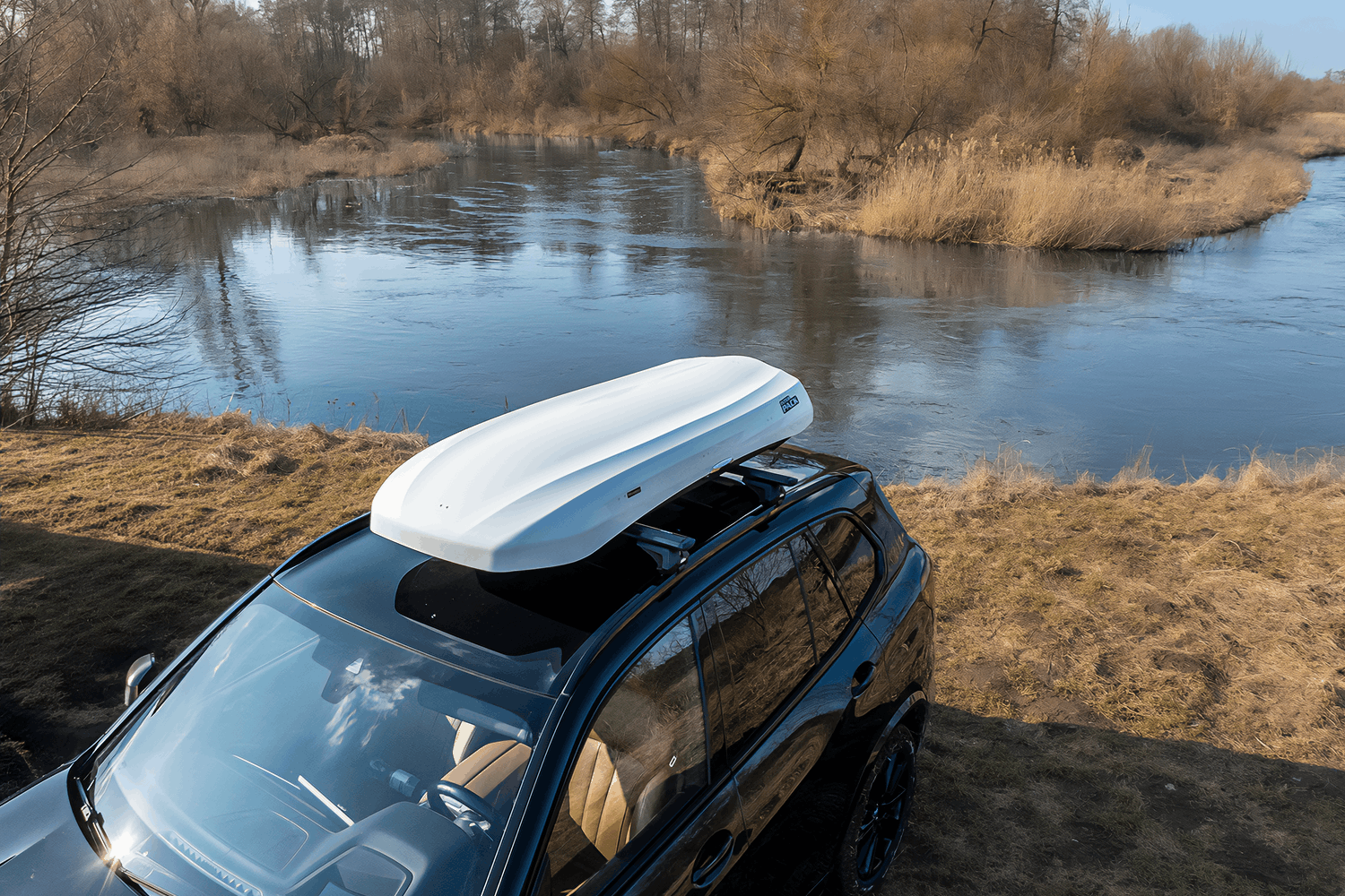  Biały, niski i nowoczesny box dachowy zamontowany na czarnym SUVie marki BMW. Łąka z wąską rzeką w tle. Na drugim brzegu rzeki widoczny gęsty las w żółtej kolorystyce.