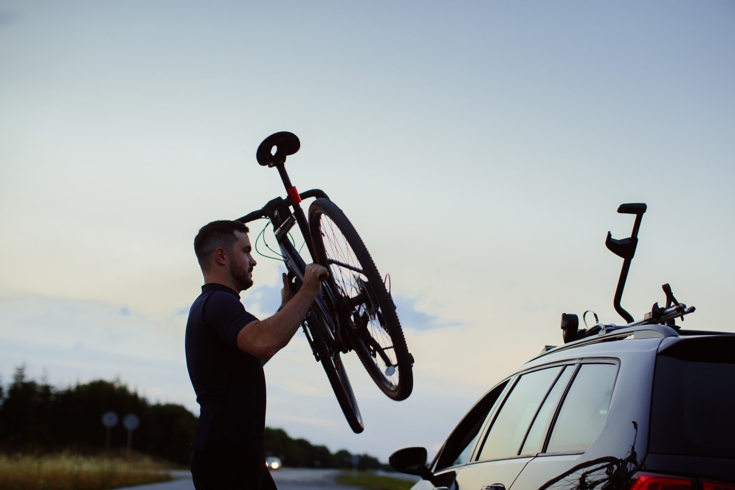 Wieczór. Mężczyzna podnoszący rower by umieścić go w uchwycie rowerowym widocznym na dachu auta. 